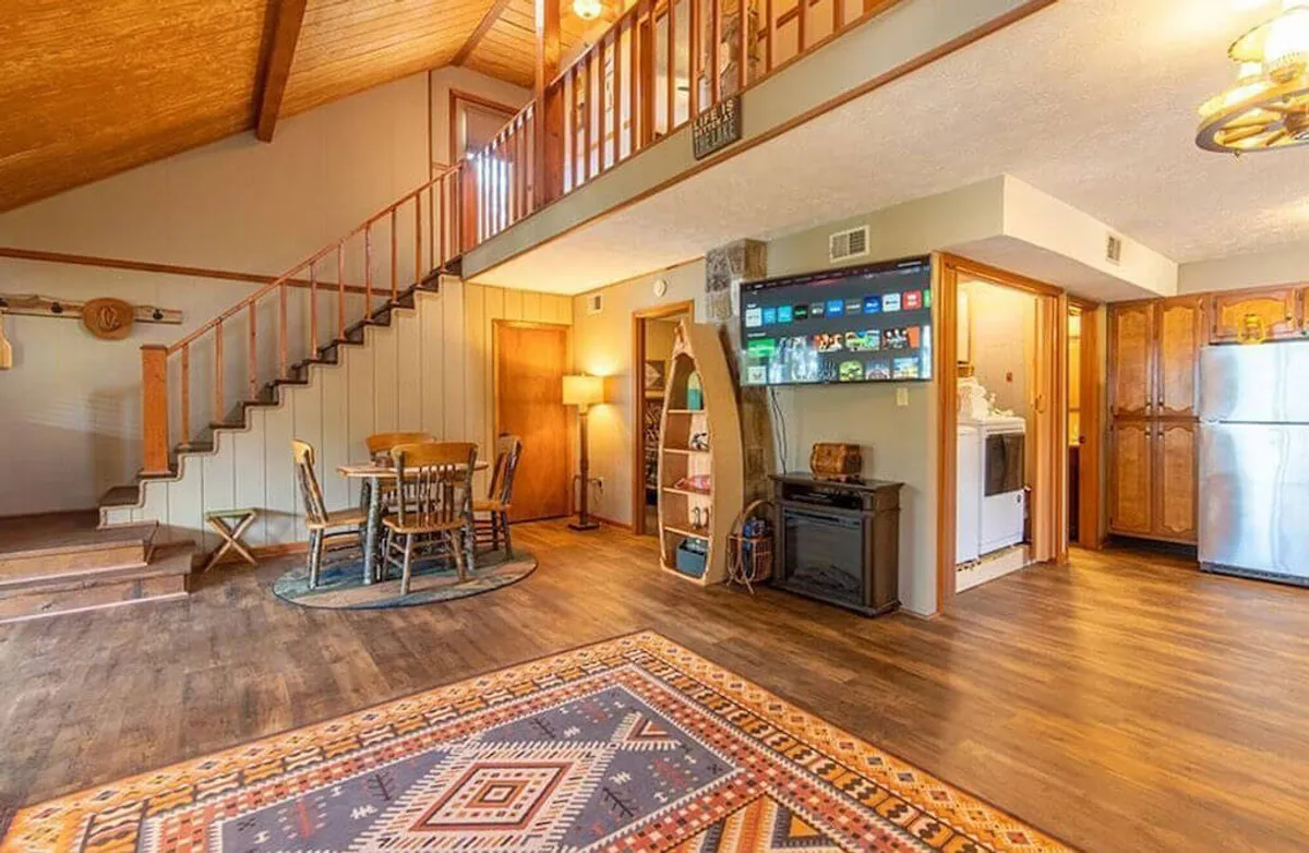This image features a welcoming interior view of the cottage living area, including a staircase, dining table, area rug, and a television unit. Natural light fills the space, creating a cozy and inviting atmosphere that is ideal for working remotely or enjoying downtime. The scene hints at a space where guests can set up their laptop, work comfortably, and feel connected to the relaxing ambiance of the cottage.