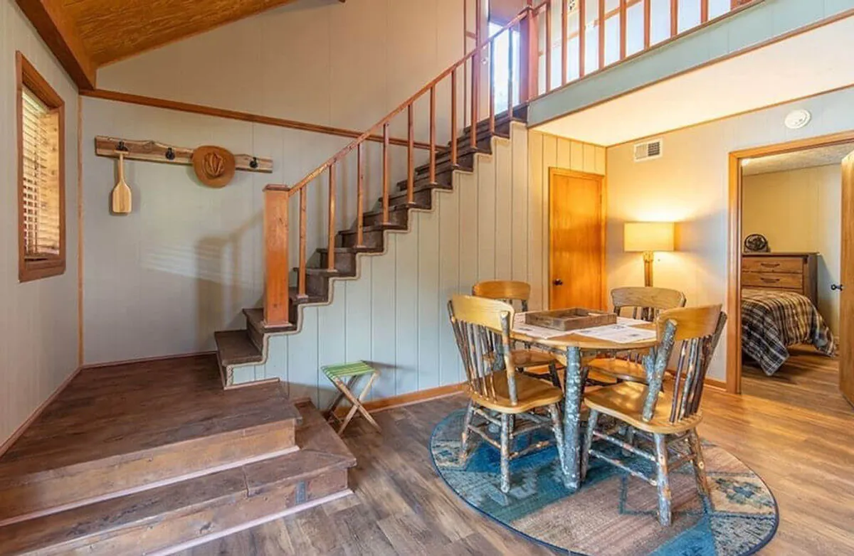 This inviting interior image captures a cozy dining area with a prominent wooden table beneath a stylish light fixture, adjacent to a staircase. The setup suggests a space where people gather for family meals, game nights, or relaxed conversations.