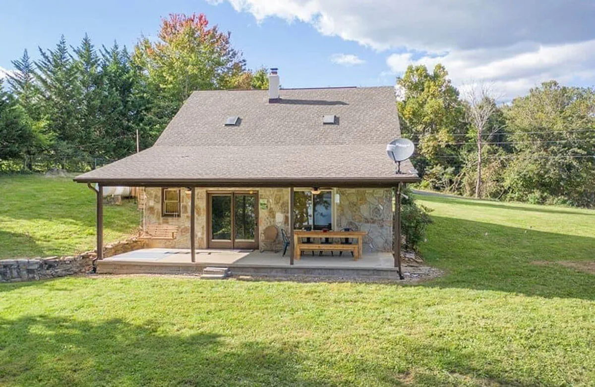 An exterior image of a patio area with a sturdy wooden dining table set on a lush green lawn surrounded by trees. The scene is tranquil, offering a peaceful space for outdoor dining, relaxation, and enjoying the natural surroundings.