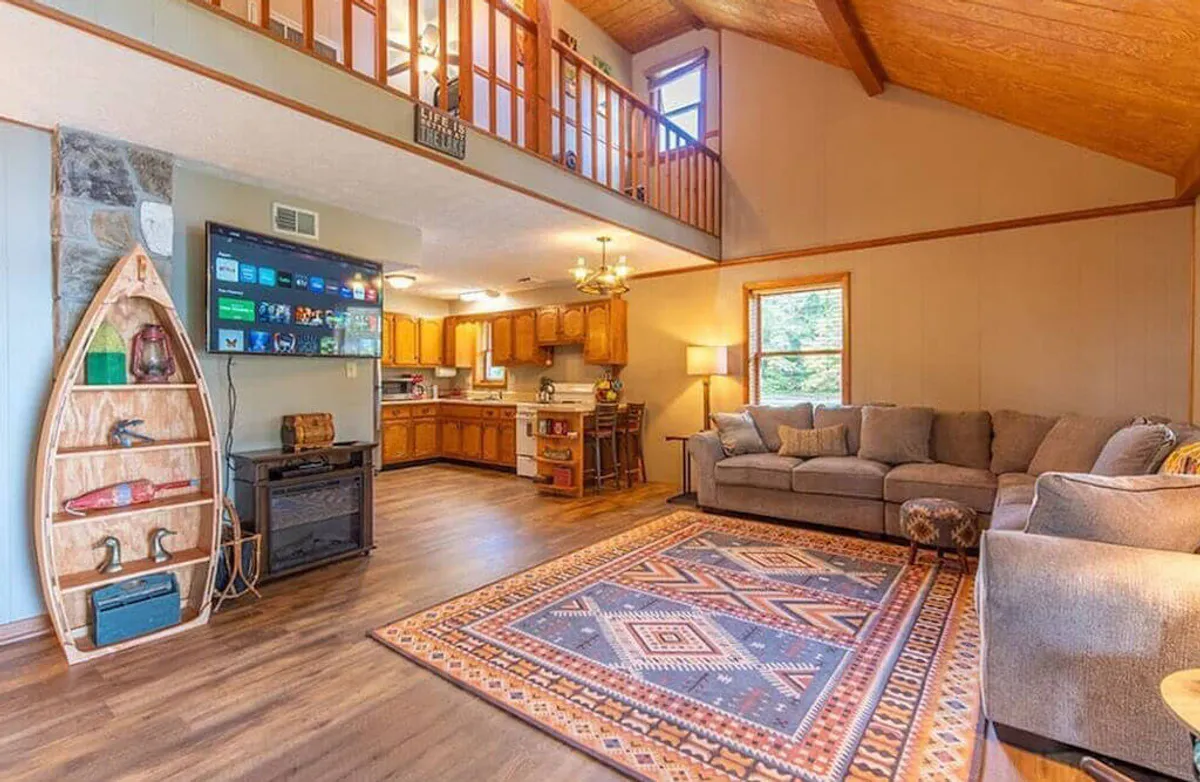 A spacious living room featuring a large sectional sofa arranged around a colorful area rug, a stone feature wall adorned with wooden boat decor, and a modern TV unit. The area is carefully staged for relaxation and gatherings, with ample seating and thoughtful design touches that evoke coziness and warmth.