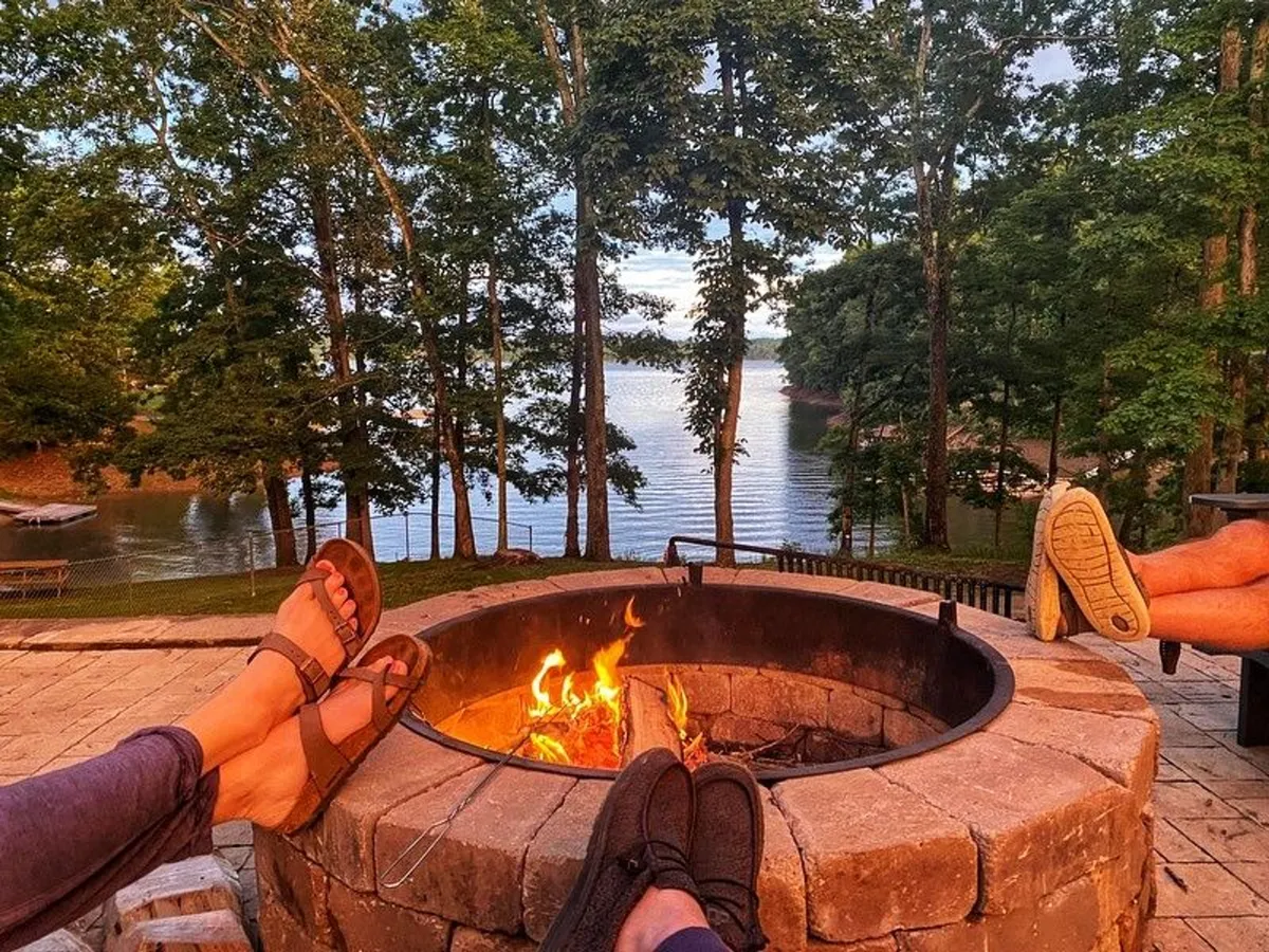 A cozy fire pit scene with families gathered around, overlooking the lake and framed by trees. The image communicates communal outdoor enjoyment—storytelling, s’mores, and bonding—particularly in the evening. The presence of the lake and picnic area links directly to the section’s callouts of backyard barbecues and gathering spots.