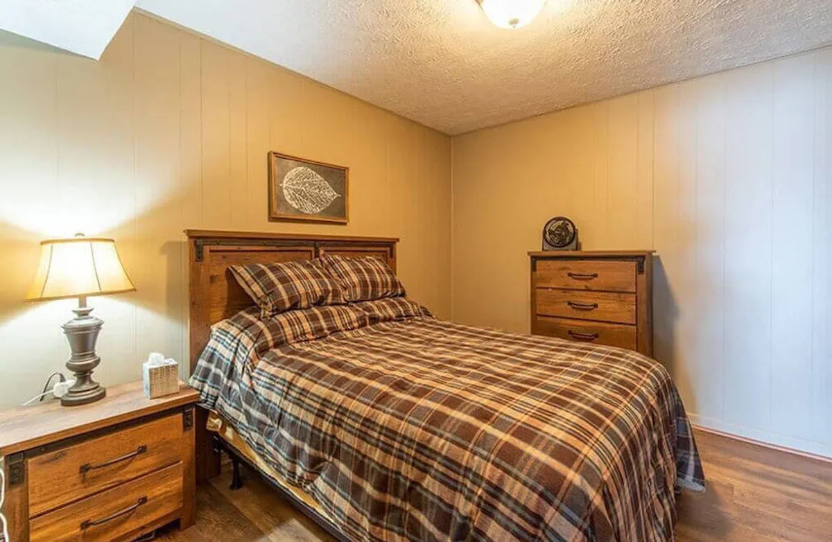 A cozy queen bedroom featuring a bed with plaid bedding, a nightstand with a lamp, a wooden dresser, and framed artwork on the wall. Sunlight filters softly through the window, creating a comfortable and restful atmosphere ideal for unwinding.