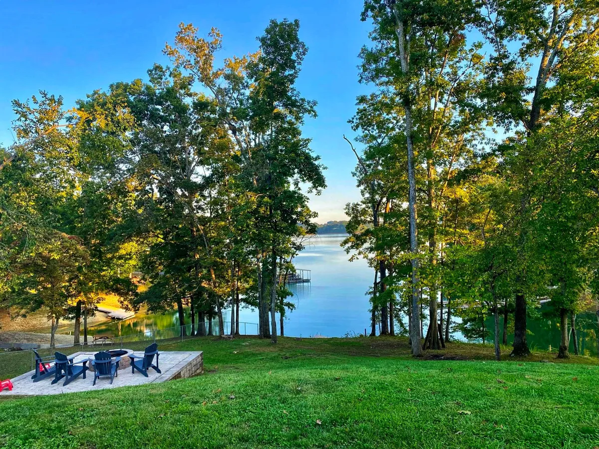 The image showcases the tranquil exterior of a lakefront cottage framed by lush trees, with a private patio overlooking serene waters. A fire pit sits invitingly near the shoreline, hinting at cozy evenings, while the spacious outdoor area offers perfect spots for gathering, relaxing, or enjoying sunrise views over the lake.