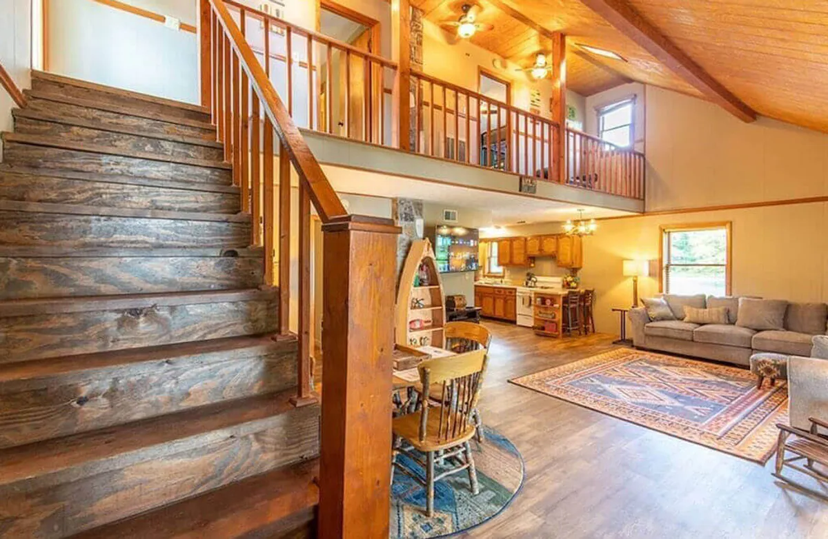 A cozy open-concept layout combining a dining table, kitchen area, and comfortable living room seating. The staircase and area rug provide visual interest, while large windows offer glimpses of the outdoors. The space is filled with natural light and centers on communal living.