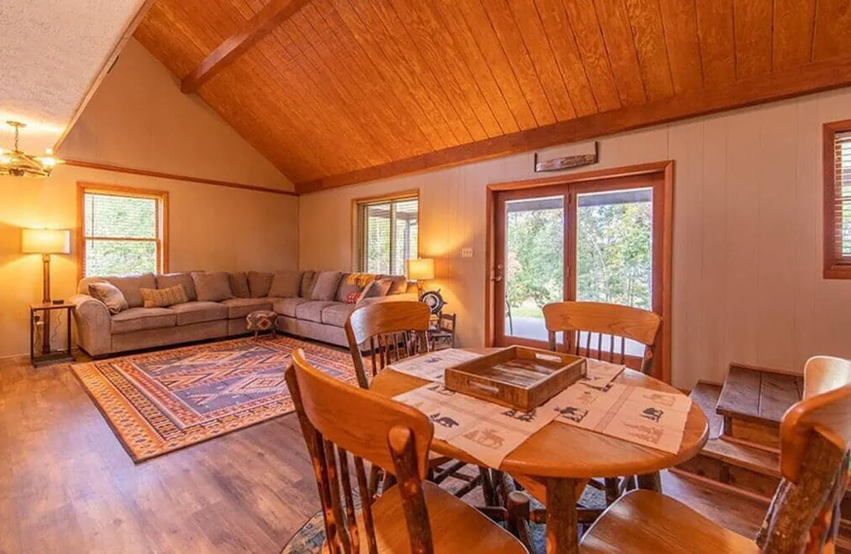 An open-plan interior showing both a comfortable sofa and a welcoming dining table set atop a patterned rug, with large windows allowing natural light to flood the room. The space feels bright and cheerful, ideal for shared meals or relaxed conversations.