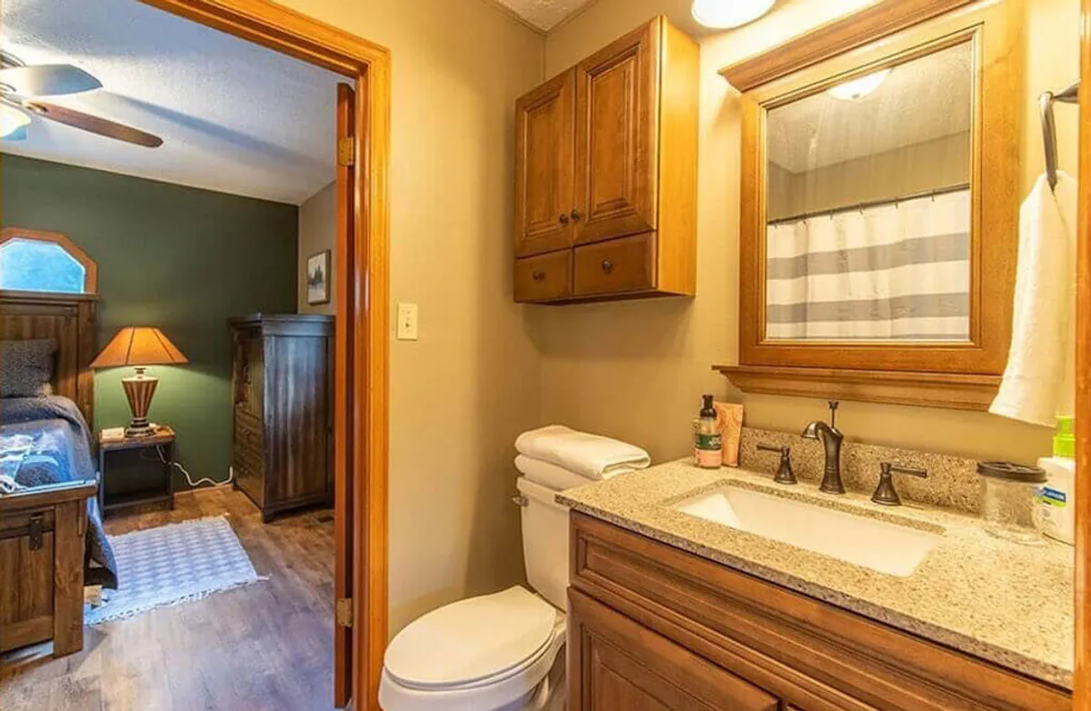 A serene bathroom featuring warm wooden cabinetry, a granite countertop, a large framed mirror, and neatly stacked fresh towels. Morning light streams through the window, highlighting the space’s calm and welcoming ambiance.